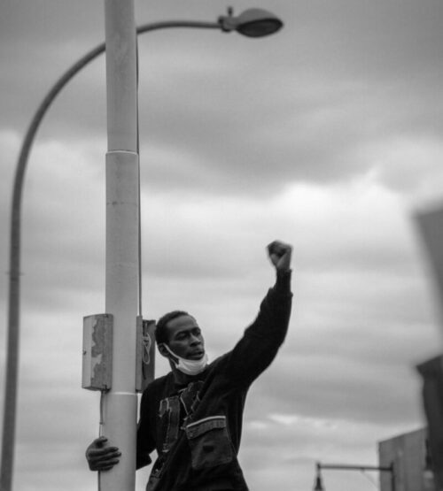 Black and white photograph of a man on a street lamp, protesting during Black Lives Matter protests in Brooklyn, New York.