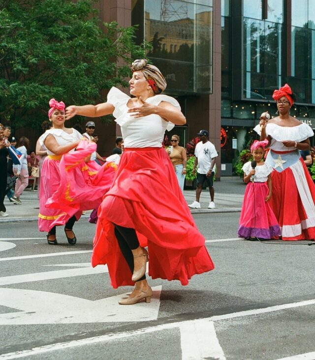 Photograph of women dancing on the street in traditional costume with red ruffled skirts and white blouses.