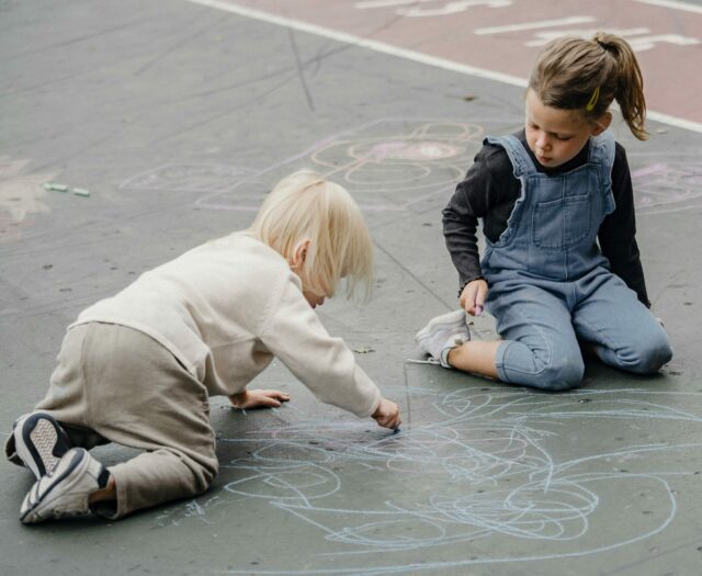 Two young children drawing on the ground with chalk.