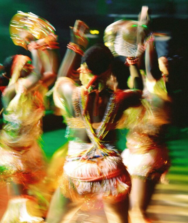 Blurred, highly saturated color photograph of women dancing in a circle with their hands aloft.